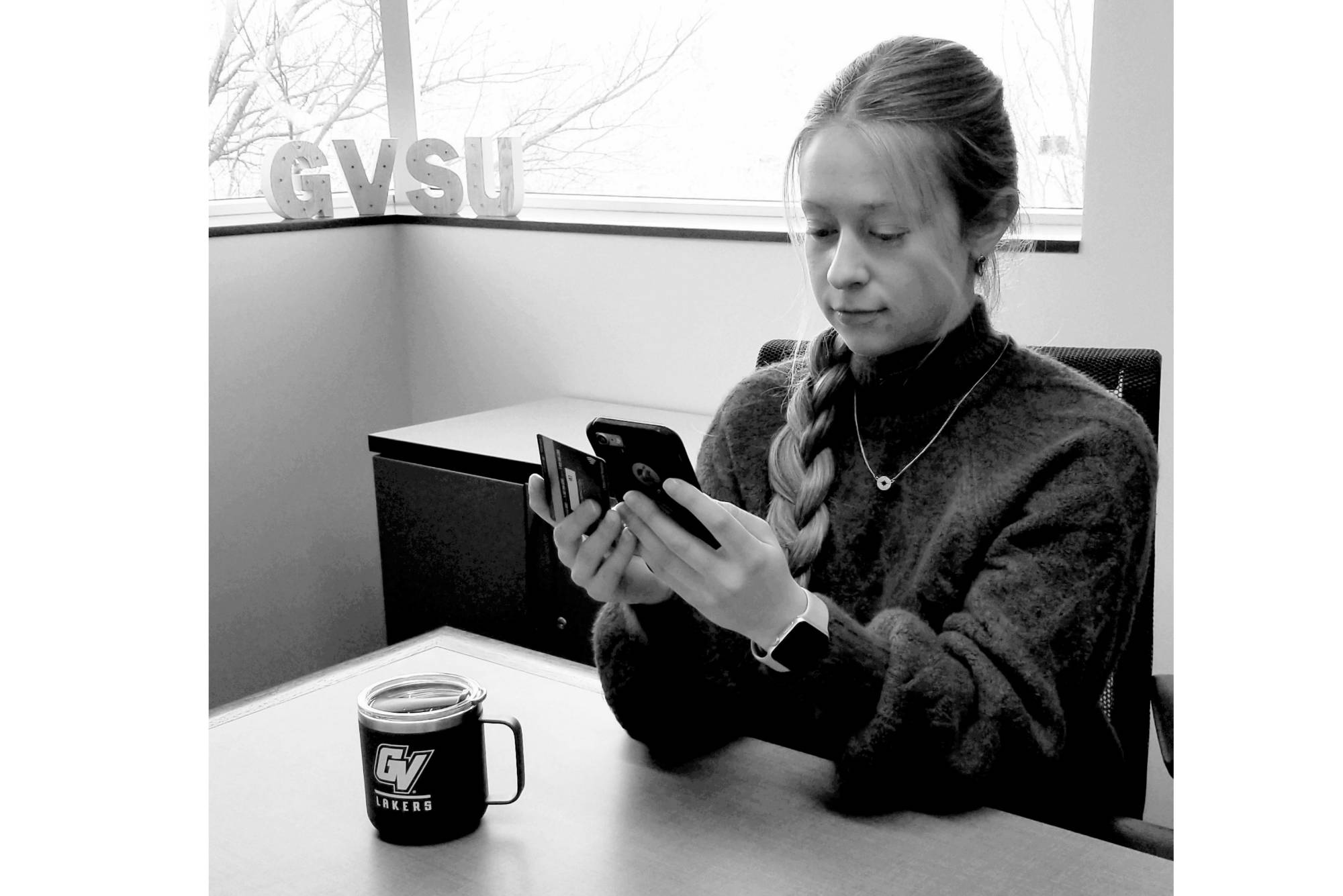 GVSU Student sitting at desk doing a cashless transaction on mobile phone with GVSU coffee mug on desk
